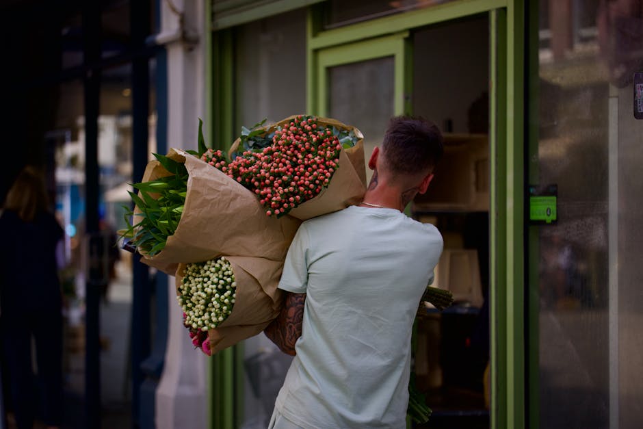 A young man with short brown hair, wearing a white t-shirt, is holding a large bouquet of fresh flowers wrapped in brown paper outside a flower shop with a lime green door frame. The bouquet features