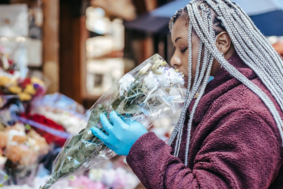 A woman with braided, silver-colored hair and wearing a maroon fleece jacket and bright blue disposable gloves is carefully holding a large bouquet of white roses wrapped in clear plastic, inspecting 