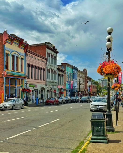 A colorful street scene featuring a row of historic Victorian-style buildings with decorative facades in pastel shades of pink, blue, and cream. The sky above is partly cloudy with patches of blue and