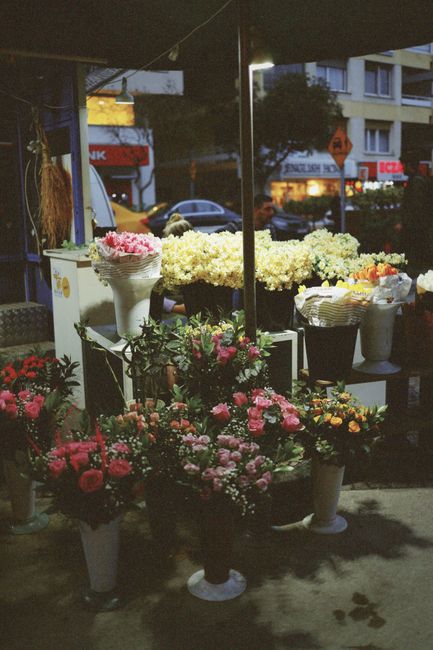 A display of fresh floral arrangements at an outdoor flower stall during dusk, featuring bouquets of pink roses, white chrysanthemums, and mixed seasonal flowers arranged in white, black, and terracot