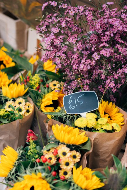 A vibrant floral display featuring handmade bouquets arranged with bright yellow sunflowers, small yellow and white daisies, and red accent flowers, all wrapped in brown paper. In the background, a la