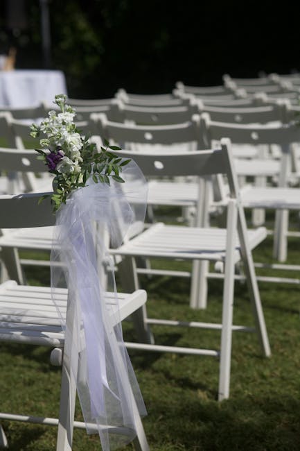 A wedding ceremony setup featuring a white wooden chair decorated with a small floral arrangement of white and purple flowers, complemented by green foliage, tied with a soft white tulle ribbon. The a
