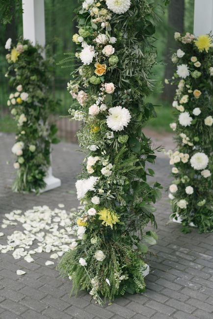 A tall, cylindrical floral arrangement featuring a lush mix of white and light yellow flowers, including chrysanthemums and roses, complemented by abundant green foliage. The arrangement is displayed