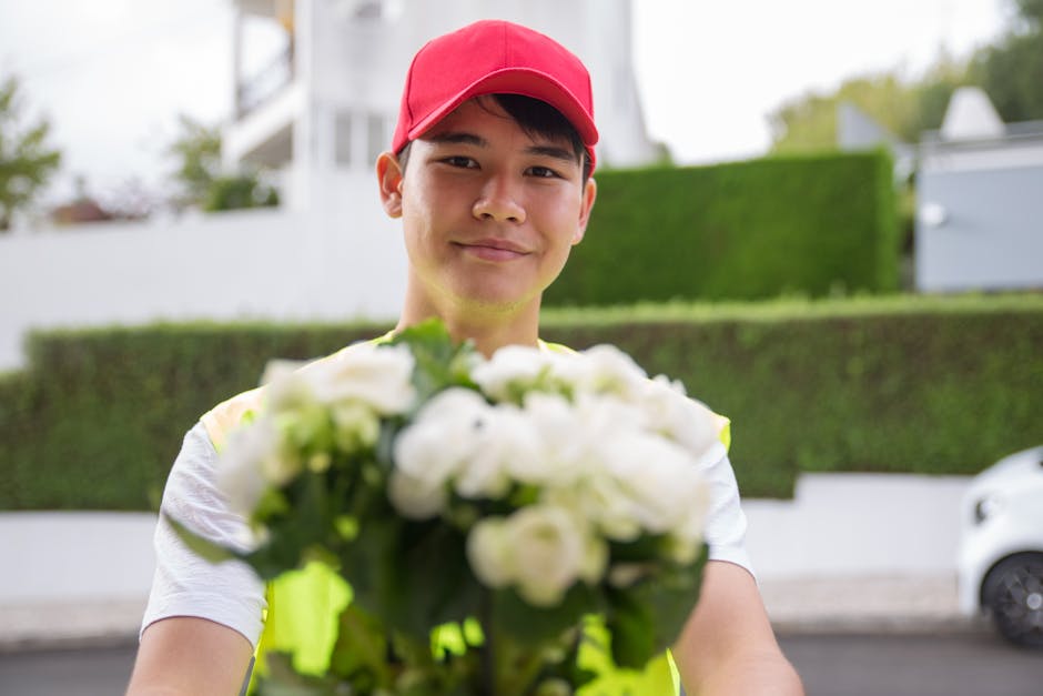 A delivery person wearing a red cap and a high-visibility vest holds out a bouquet of fresh white flowers with green leaves towards the camera. The bouquet is composed of tightly packed blossoms, poss