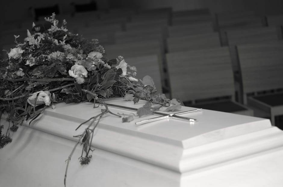 A white coffin is decorated with a large, lush floral arrangement composed of various flowers and greenery, including white blossoms and leafy foliage. The flowers are arranged in a cascading style, d